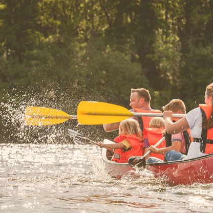 Eine fünfköpfige Familie fährt Kanu auf einem See. Die Paddel spritzen dabei mit Wasser..