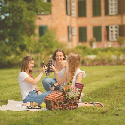 Drei Freundinnen prosten sich mit Limonade auf einer Picknickdecke vor dem Schloss Eutin zu