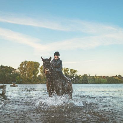 Ein Frau reitet mit ihrem Pferd durch einen See. Sie befinden sich am Ufer.