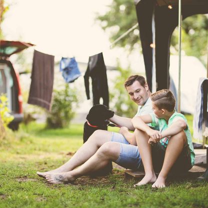 Ein Vater sitzt mit seinem Sohn und Hund vor einem Campingwagen