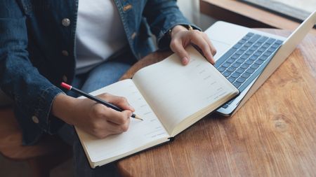 Frau schreibt in ein Notizheft, ihr Laptop liegt neben ihr. 