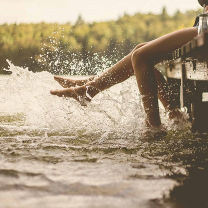 Zwei Personen sitzen auf einem Steg und schleudern mit den Füßen das Wasser eins Sees hoch.