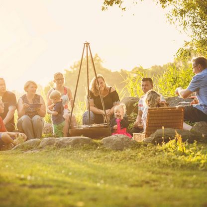 Eine Familie sitzt rund um ein Lagerfeuer, gleich neben einer Badestelle.